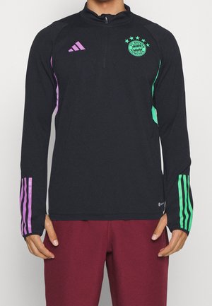Osoba ubrana w czarną bluzę Adidas z długim rękawem z logo FC Bayern Monachium, fioletowe paski na lewym rękawie, zielone paski na prawym rękawie oraz bordowe spodnie.