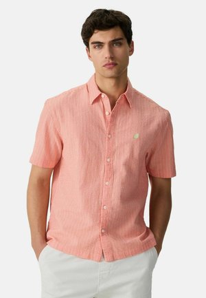 Mann trägt ein korallenfarbenes, kurzärmeliges Hemd mit Knöpfen und einem kleinen Limetten-Patch auf der Brust, kombiniert mit weißen Hosen, Hände in den Taschen.