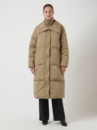 Cappotto lungo imbottito beige con colletto alto e chiusura frontale a bottoni; presenta cuciture diagonali e una vestibilità comoda. Indossato sopra pantaloni neri.