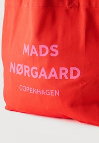 Φωτεινό κόκκινο ύφασμα με ρόζ κείμενο που γράφει "MADS NØRGAARD COPENHAGEN" τυπωμένο στην επιφάνεια.