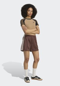 Calções de desporto castanhos com riscas brancas nas laterais, combinados com uma blusa de manga curta listrada em bege e preto, e sapatilhas pretas com um detalhe em tom de terra.