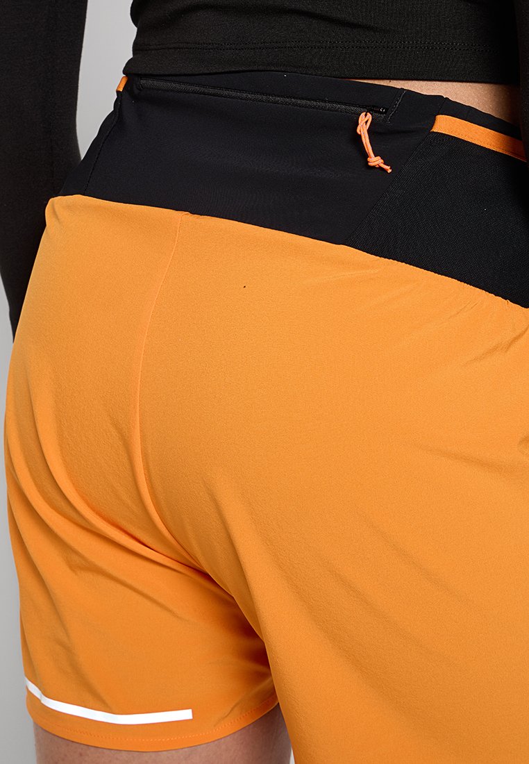 Pantalones cortos deportivos naranjas con una cintura negra, que presentan un bolsillo lateral y un detalle en blanco. La tela parece suave y flexible.