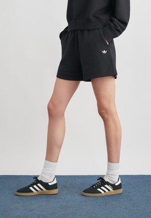 Osoba oblečená v čiernych šortkách Adidas a čiernych teniskách Adidas s bielymi pruhmi a bielymi ponožkami, stojaca na modrej podlahe.