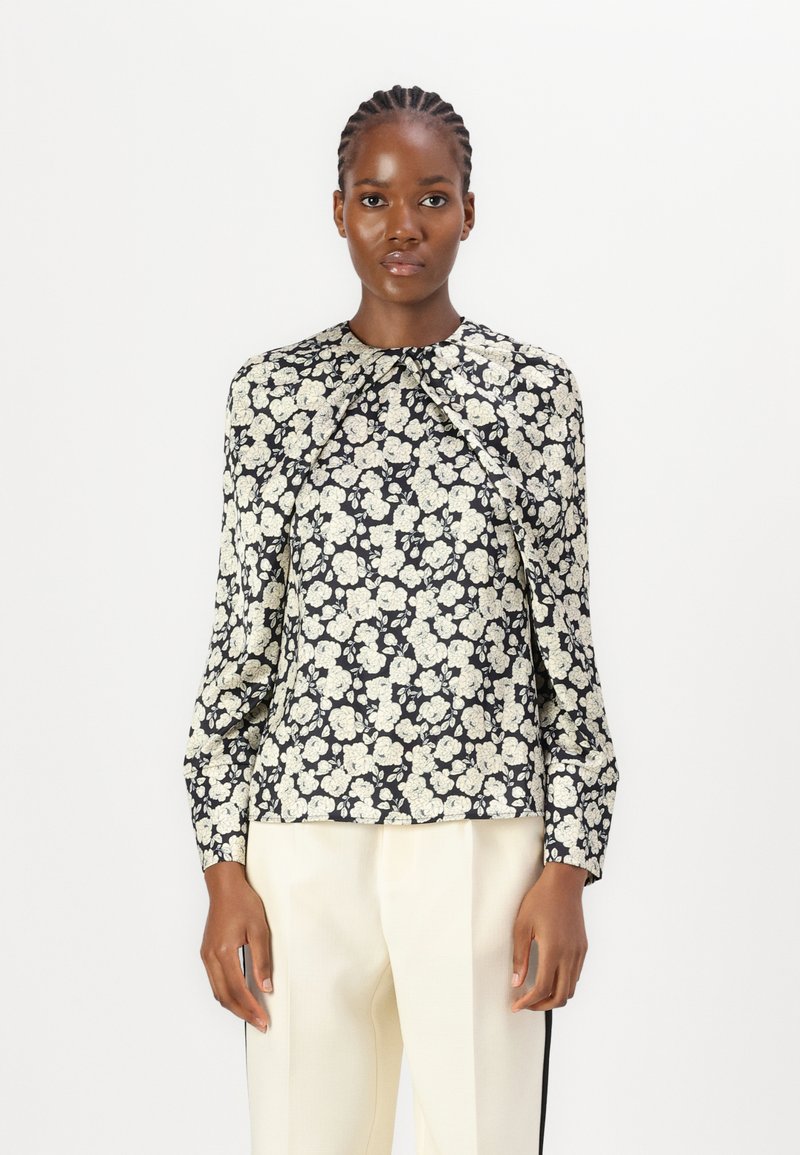Langärmlige Bluse aus schwarzem Stoff mit einem cremefarbenen Blumenmuster. Verfügt über einen gerafften Ausschnitt und einen leicht lockeren Schnitt. Kombiniert mit cremefarbenen Hosen.