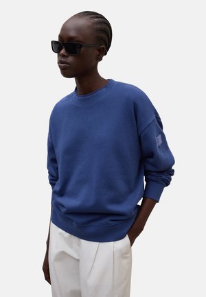 Persoon met een blauwe lange mouwen trui, witte broek, zwarte zonnebril en gevlochten haar, staand tegen een effen lichte achtergrond.