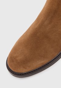 Bruine suède enkelboot met een ronde neus, donkerbruine rubberen zool en gedetailleerde stiksels langs de rand, met een soepele textuur.