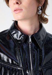 Fekete műbőr dzseki, cipzáros zárással, nagy gallérral és texturált felülettel, látható varrással és modern siluettel.