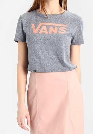 Nainen pukeutuneena harmaaseen Vansin t-paitaan, jossa on vaaleanpunainen logo, ja korkeavyötäröiseen vaaleanpunaiseen hameeseen valkoisella taustalla.