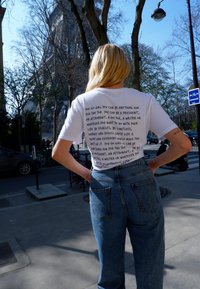 Witte t-shirt met zwarte tekst op de achterkant met motiverende zinnen; gecombineerd met hoog-ruggedragen blauwe spijkerbroek. Casual pasvorm, korte mouwen.