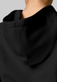 Nahaufnahme eines schwarzen Kapuzenkleidungsstücks, das auf dem oberen Rücken und Nacken einer Person getragen wird, vor einem einfarbigen grauen Hintergrund.