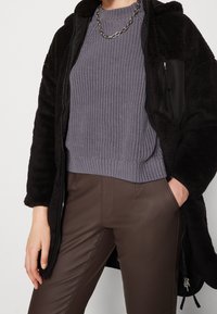 Abrigo negro de pelaje sobre un suéter de punto texturizado gris claro y pantalones ajustados marrones. Incluye un collar de cadena plateado y detalles con cremallera.