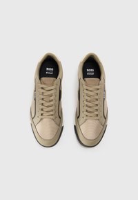Beige treningsko med overdel i mesh og syntetisk materiale, flate lisser og dempende såle; har svarte og hvite detaljer.