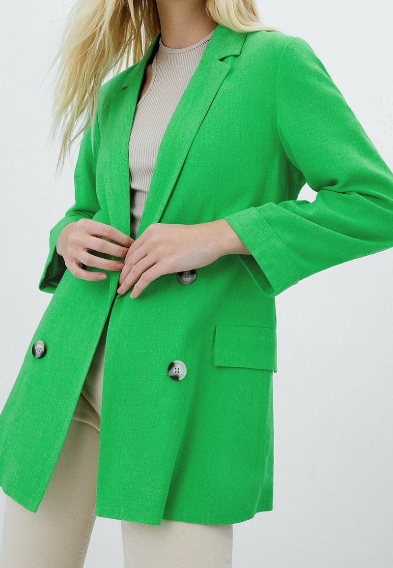 Femme boutonnant un blazer croisé vert vif porté sur un haut beige et un pantalon sur un fond uni.