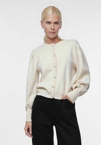 Crème kleurige cardigan met ronde halslijn en zes grote knopen. Heeft opgezette mouwen en geribbelde manchetten, gecombineerd met zwarte broek.
