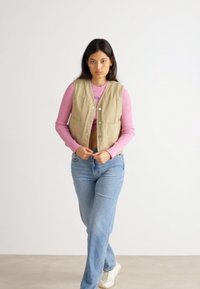 Beige polstret vest med frontlommer over en rosa ribbet longsleeve skjorte, sammen med lyseblå jeans og hvite joggesko.