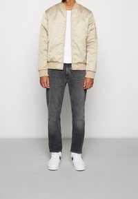 Giacca bomber satin beige con chiusura a zip, polsini a coste e taschino, abbinata a jeans slim-fit grigi scuri e sneakers bianche.