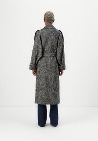 Cappotto lungo in lana grigia a zigzag con vestibilità rilassata, maniche ampie e vita con cinturino, indossato sopra jeans blu a zampa.