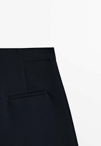 Gros plan sur la taille d'un pantalon noir sur mesure avec une poche arrière horizontale visible sur un fond blanc uni.