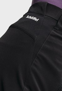 Pantalon d'extérieur noir ajusté avec couture renforcée et logo blanc Schöffel près de la taille sur une personne portant du violet.