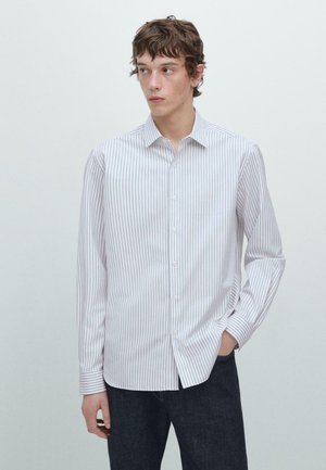 Junger Mann, der ein weißes Hemd mit dünnen vertikalen Streifen und dunkle Hosen trägt, mit einer Hand in der Tasche stehend und nach links blickend.