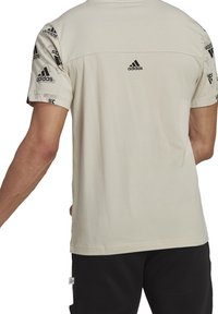 Lyhythihainen, vaaleanbeige Adidas-t-paita, jossa musta logo selässä ja olkapäässä, valmistettu pehmeästä, sileästä puuvillakankaasta.