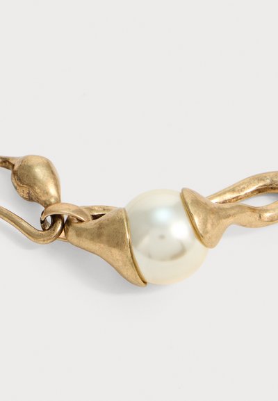Collier en métal doré avec une grande perle blanche centrale maintenue par deux fermoirs métalliques incurvés sur fond blanc.
