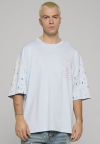 Jasnoniebieska, oversizeowa koszulka z krótkimi rękawami. Rękawy ozdobione kolorowymi wzorami przypominającymi konfetti. Z przodu znajduje się prosty wzór graficzny. Wykonana z bawełny.