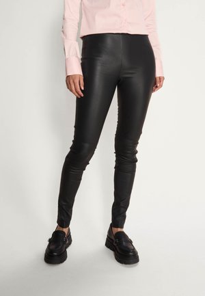 Person trägt schwarze eng anliegende Lederhose, schwarze klobige Loafers und ein hellrosa langärmliges Hemd mit Knöpfen vor einem schlichten Hintergrund.