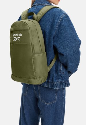 Persona con zaino Reebok verde oliva e giacca di jeans e jeans, vista da dietro su sfondo bianco.