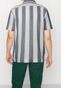 Chemise à manches courtes rayée gris clair et bleu marine, en tissu texturé. Comprend un col et un ourlet droit. Pantalon vert en dessous.