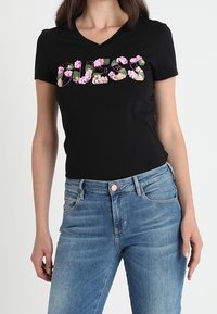 Musta v-pääntie t-paita, jossa on sana "GUESS" kukkakuosilla; yhdistetty sinisiin farkkuihin. Kangas on sileä ja siinä on slim fit -muotoilu.