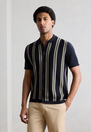 Άνδρας που φοράει μπλε σκούρο κοντομάνικο πουκάμισο με κάθετες μπεζ ρίγες και μπεζ παντελόνι, στέκεται μπροστά σε απλή φωτεινή τοίχο.