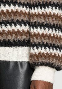 Den stickade tröjan har bruna, svarta och vita zigzag-mönster, med en ribbad krämfärgad mudd. En svart läderyta är delvis synlig nedanför.