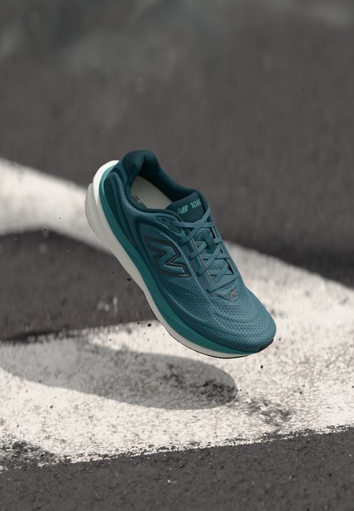 Modrá běžecká bota New Balance se šňůrkami, která visí nad asfaltovou cestou s bílými malovanými značkami a rozptýleným drobným štěrkem.
