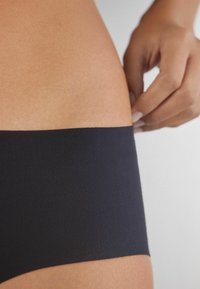 Zwarte naadloze hipsteronderbroek met een soepele textuur, voorzien van een brede tailleband, gedeeltelijk getoond tegen een huidskleurige achtergrond.