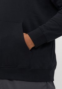 Hand inside the front pocket of a black sweatshirt worn with dark gray pants, showing sleeve cuff and waistband details.