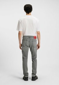 Grå denim jeans med et rødt logo-patch bagpå, parret med en ensfarvet hvid T-shirt, set fra背en mod en ensfarvet baggrund.