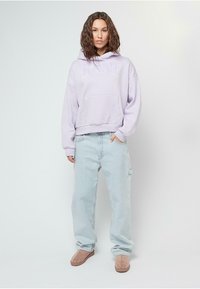 Lavendelfarbene übergroße Kapuzenjacke mit frontaler Tasche und Logo, kombiniert mit hellblauen, lockeren Jeans und beigen Slippern. Das Model steht vor einem neutralen Hintergrund.
