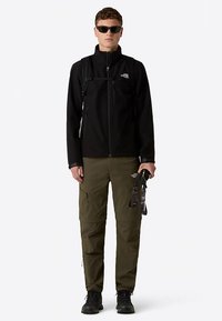 Svart fleecetröja med dragkedja fram, gröna cargobyxor, bär solglasögon, håller i en rem, vattentäta stövlar och en ryggsäck.