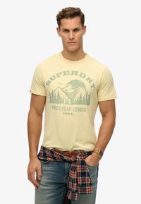 Gul bomulds T-shirt med et bjergmotiv i grønt, rund halsudskæring, korte ærmer, parret med blå jeans og en ternet skjorte bundet om livet.
