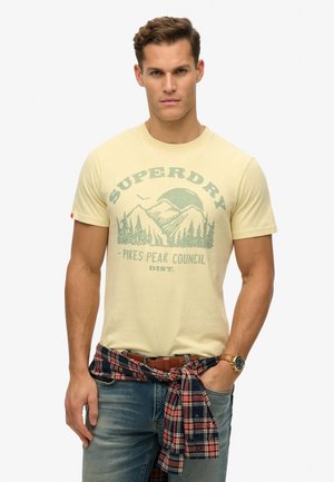 Yellow cotton T-shirt featuring a mountain graphic in green, round neckline, short sleeves, paired with blue jeans and a plaid shirt tied at the waist.