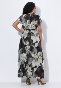 Vrouw met lang, krullend haar, gekleed in een zwarte bloemenmaxi-jurk die op de taille is vastgebonden en gouden hoge hakken, staand tegen een witte muur.
