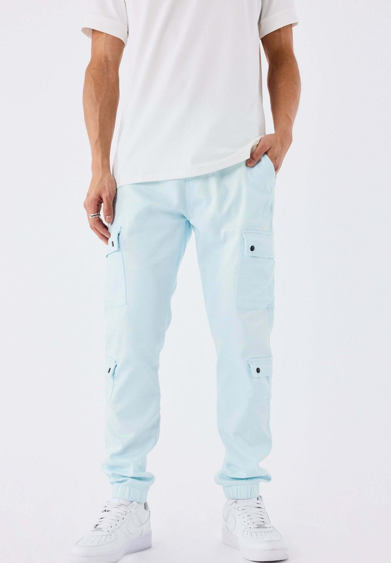 Persona che indossa pantaloni cargo azzurri chiari con polsini elastici e sneakers bianche, con le mani in tasca, su uno sfondo bianco semplice.