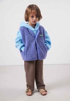 Mlad otrok z rjavimi lasmi v svetlo modri kapucarji, vijolični flis telovnik, rjavih hlačah in bež sandalih, stoji ob belem ozadju.