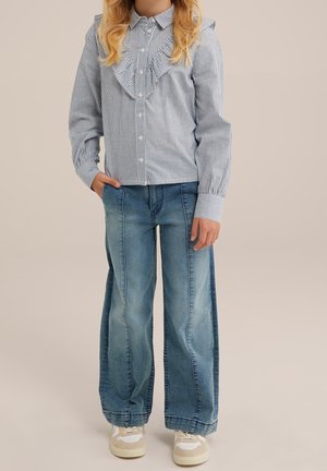 Dziecko ubrane w niebiesko-białą koszulę w paski z falbankami i szerokie niebieskie dżinsy, stojące z jedną ręką w kieszeni.