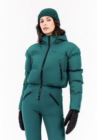 Teal geïsoleerde pufferjack met een ritssluiting aan de voorkant en zakken, gecombineerd met bijpassende broek. Zwarte handschoenen en een geribde teal beanie maken de look af.