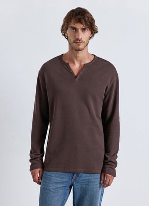Uomo con capelli corti castani che indossa una maglietta henley a maniche lunghe marrone scuro e jeans blu, in piedi davanti a uno sfondo chiaro e semplice.
