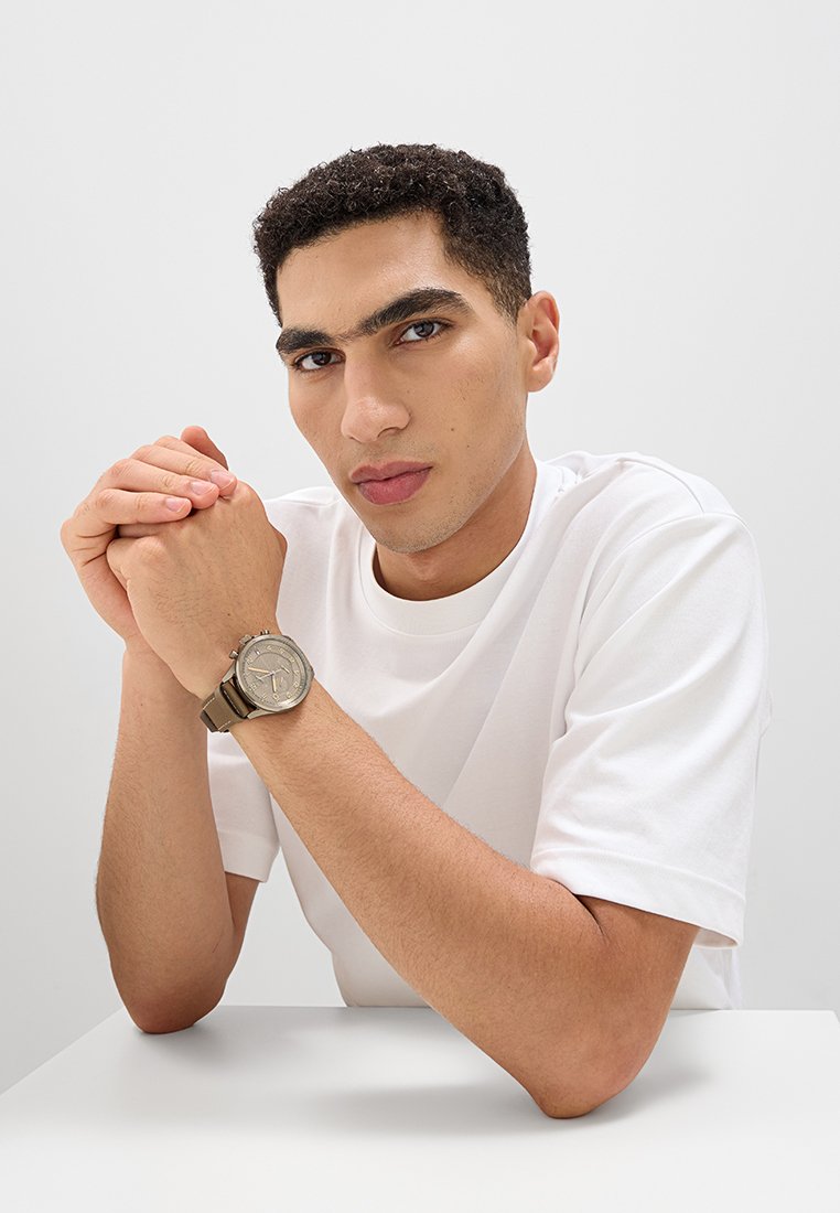 Brązowy skórzany zegarek z okrągłą srebrną tarczą, na której znajdują się proste oznaczenia godzin. Noszony na nadgarstku z krzyżującymi się rękami na białym stole.