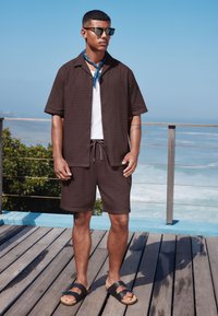 Krótka koszula z ciemnobrązowym wzorem, biały podkoszulek, pasujące spodenki, czarne sandały i niebieska chusta, na tle nadmorskiego krajobrazu.