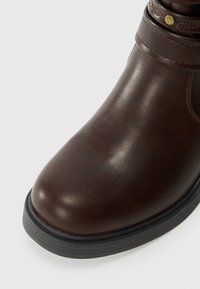 Dark brown leather ankle boot with black rubber sole and decorative strap with brass stud near the top.